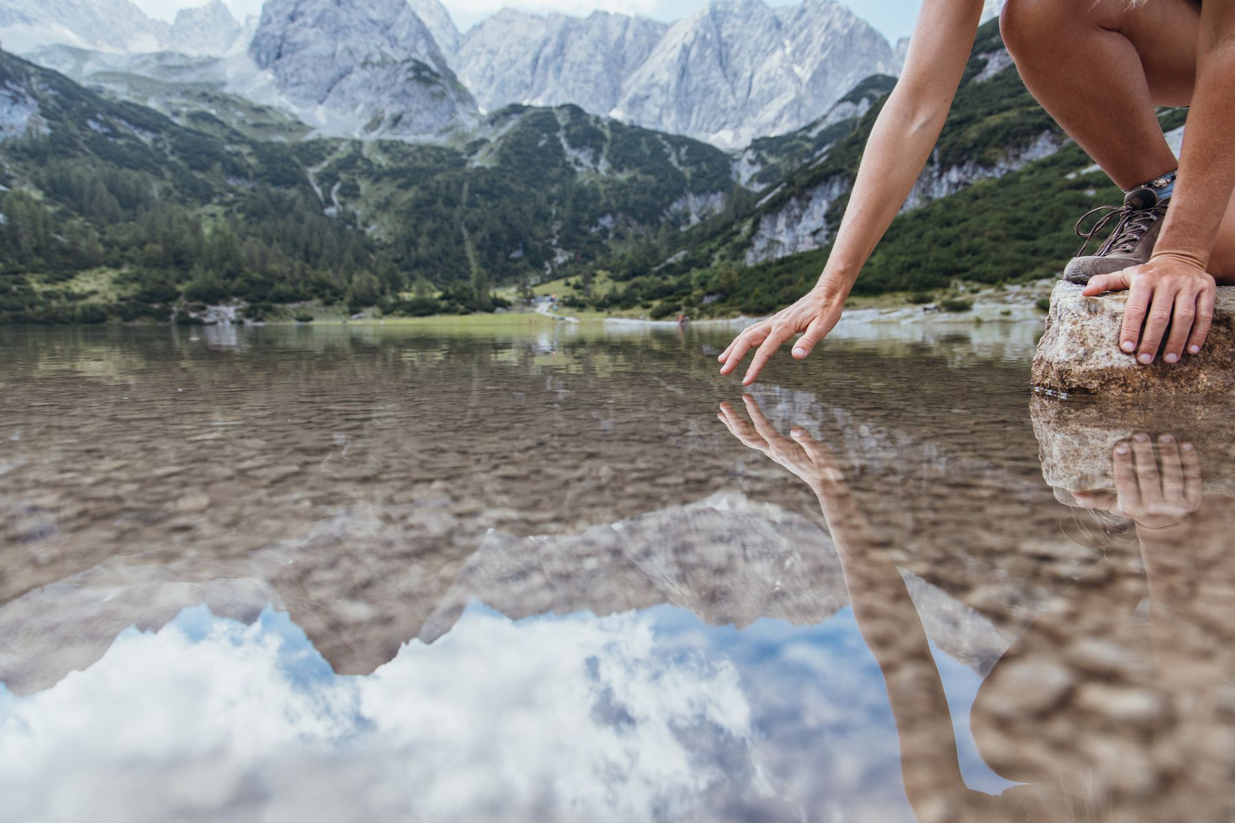 Klarer Bergsee vor Berggipfeln, eine Hand berührt das Wasser